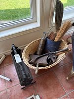 Full view of the wicker basket filled with fireplace tools including wooden broom brushes in a black metal container, a metal oil canister, and a large metal shovel with wooden handle. The black wrought iron wood holder placed on the floor next to the basket.