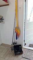 Photo showing three fishing rods standing upright with reels, a folded colorful hammock hanging on the wall, and a black tackle box open on the floor containing fishing accessories.