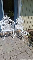 Two white cast iron garden chairs and one matching plant stand positioned on outdoor stone patio against building wall and glass door.