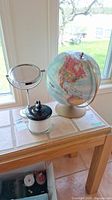 Photo showing the globe, vanity mirror, and lidded jar filled with bath salts arranged on a wooden surface near a window.