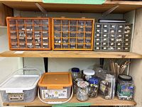 Two wooden shelves have three orange plastic organizer cases with multiple small transparent drawers holding assorted screws, bolts, nuts, and washers. Below shelves are additional plastic containers and jars with more hardware parts.