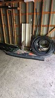 Photo of hoses stored along a shed wall, showing large coil of black PVC hose, green garden hoses, and corrugated black PVC hoses with orange fittings on the floor.