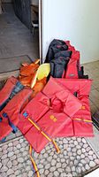 Photo showing multiple life jackets including large black and red, orange, and yellow life jackets laid on the floor.