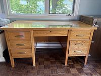 Front view of oak wood office desk with six drawers and glass top protector under window light.