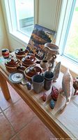 Overview of four brown onion soup bowls with lids sitting in wicker baskets, two stoneware decor pieces, metal cups, and a framed Canadian stamp collection near window.