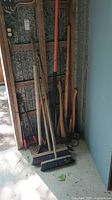 Full view showing various long-handled outdoor tools including axes, saws, brooms, tamper, and branch trimmer leaning against a wall