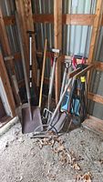 Overview of various garden and outdoor tools leaning against a wood wall, including shovels, spades, forks, and an extension cord.