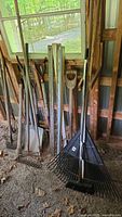 Group of outdoor garden tools leaning against a wall including shovels, rakes, pickaxe, potato forks.