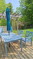 Photo of patio table with umbrella and two chairs set on wooden deck, shown assembled under pergola structure.