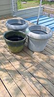 Three large plastic planters shown together on wooden deck, two light grey with wide rim and one olive green.
