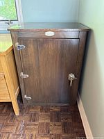 Frontal view of a dark wood vintage ice box converted cabinet with metal hinges and latch on a wooden parquet floor near a window and light wall.