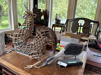 Photo showing the side view of the large woven rooster and the painted wooden duck placed on a wooden table by a window.