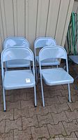 Four vintage metal folding chairs painted light blue arranged in two rows outdoors on paved surface next to a wall.