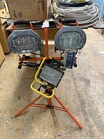 Three work lights shown: dual HDX lights on orange stand, single yellow-bordered light on tripod stand, all in industrial setting with pallets and cables around.