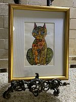 Frontal photo of the framed cat artwork with visible signature and ornate stand.