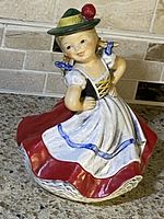 Front view of the Goebel ceramic figurine showing the girl with hands on hips in traditional white dress with red skirt and blue trims, green hat with red pompom.