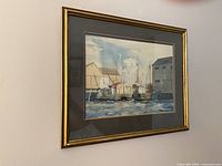 Photo showing the framed watercolor painting depicting a harbor scene with boats and buildings, hung on a wall.