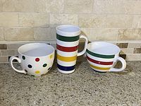 All three Hudson's Bay ceramic mugs shown side by side on countertop with light tile backsplash. Mug with polka dots on left, tall striped mug in middle, shorter striped mug on right.