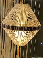 Front view of the mid-century modern Danish swag lamp showing white jute rope wrapped around to form a diamond shape with teak wood band and illuminated interior. Gold tassel hanging below.