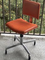 Front-right angle view of the orange 60s desk chair showing the fabric seat, leatherette bordered edges, chrome base, and wheels.
