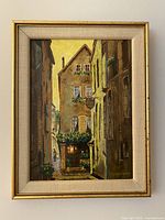 Frontal photo of oil painting on board in its original gold frame with linen matting, showing a narrow village street scene with warm tones.