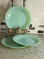 Three vintage Fire King jadeite green glass plates displayed on a counter, one held upright by an ornate metal stand, two stacked below.