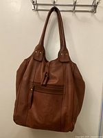 Front view of the brown leather tote bag hanging on a wall hook, showing two shoulder straps and front zippered pocket with leather zipper pull.