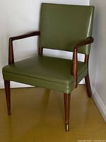 Front quarter angle view of olive green leather chair showing wood armrest and brass-tipped tapered legs.