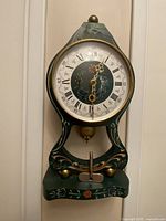 Front view of the wooden pendulum clock showing the circular clock face with Roman numerals and minute markers, decorative painted wood in green with folk art style.