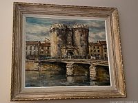 Frontal view of the oil painting showing the landscape of "Verdun" with the two towers and bridge, in a rustic white-washed wooden frame.