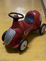 Side front angle of the red metal ride-on toy car showing black seat, steering wheel, front grille, and left wheels.