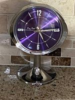Front image of the clock showing the purple dial, silver numbers, and hands on a silver metal pedestal base.