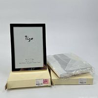 Photo showing two silver plated Tigo picture frames, one standing and one in its box wrapped in protective plastic.