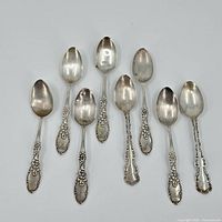 Eight ornate sterling silver spoons laid out showing bowls and decorative handles.