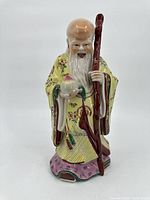 Porcelain statue of Asian elder with large forehead and white beard, wearing yellow floral robe, holding fruit and cane.
