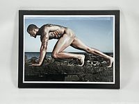 Front view of the framed color photograph of Colin Kaepernick in a natural outdoor setting, showing full body nude portrait crouched on rock with ocean background.