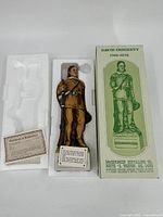 Ceramic decanter of David Crockett holding a rifle, stylized as a whiskey bottle, in original white Styrofoam packaging alongside box and certificate.