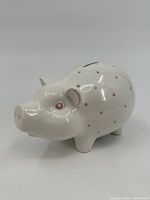 Front and side view of white ceramic piggy bank shaped as a pig with pink polka dots and eyes, showing coin slot on top.