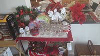Photo showing Christmas glasses with gold rims, mugs, decorative tins, poinsettias, table linens, and figurines arranged on a table.