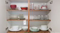 Interior cupboard shot showing shelves with assorted plates, bowls, glassware and cups neatly arranged.
