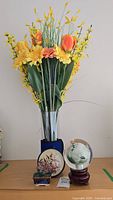 Artificial floral arrangement in glass vase along with glass globe, bonsai and framed floral art on surface