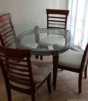 View of round clear glass top dining table with wooden legs and metal supports, surrounded by chairs.
