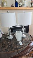 Two chrome base table lamps with white shades and a small frosted glass lamp on a glass tabletop.