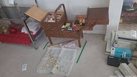 Wide view of wooden sewing box open showing multiple tiers filled with sewing notions including threads and tools. The box stands on four legs and has a handle on top.