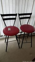 Two metal stools with red upholstered seats and black metal backs and legs placed on carpet in front of vertical blinds.