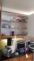 Photo showing kitchen counter with small appliances including toaster oven, toaster, coffee maker, blender base and items on upper shelves: plates, glasses, and baking sheets