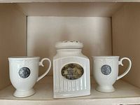 Photo of the full tea set showing two white ceramic tea cups and one tea canister with metal plaques.