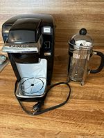 Full view of Keurig coffee maker and French press on wooden surface, showing power cord and details.