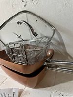 Two copper-colored pans stacked, metal fry basket on top, and clear glass lid branded Copper Chef leaning behind.