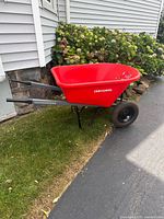 Full side-view of bright red Craftsman wheelbarrow with black steel handles and two large wheels, set on grass and pavement next to house.